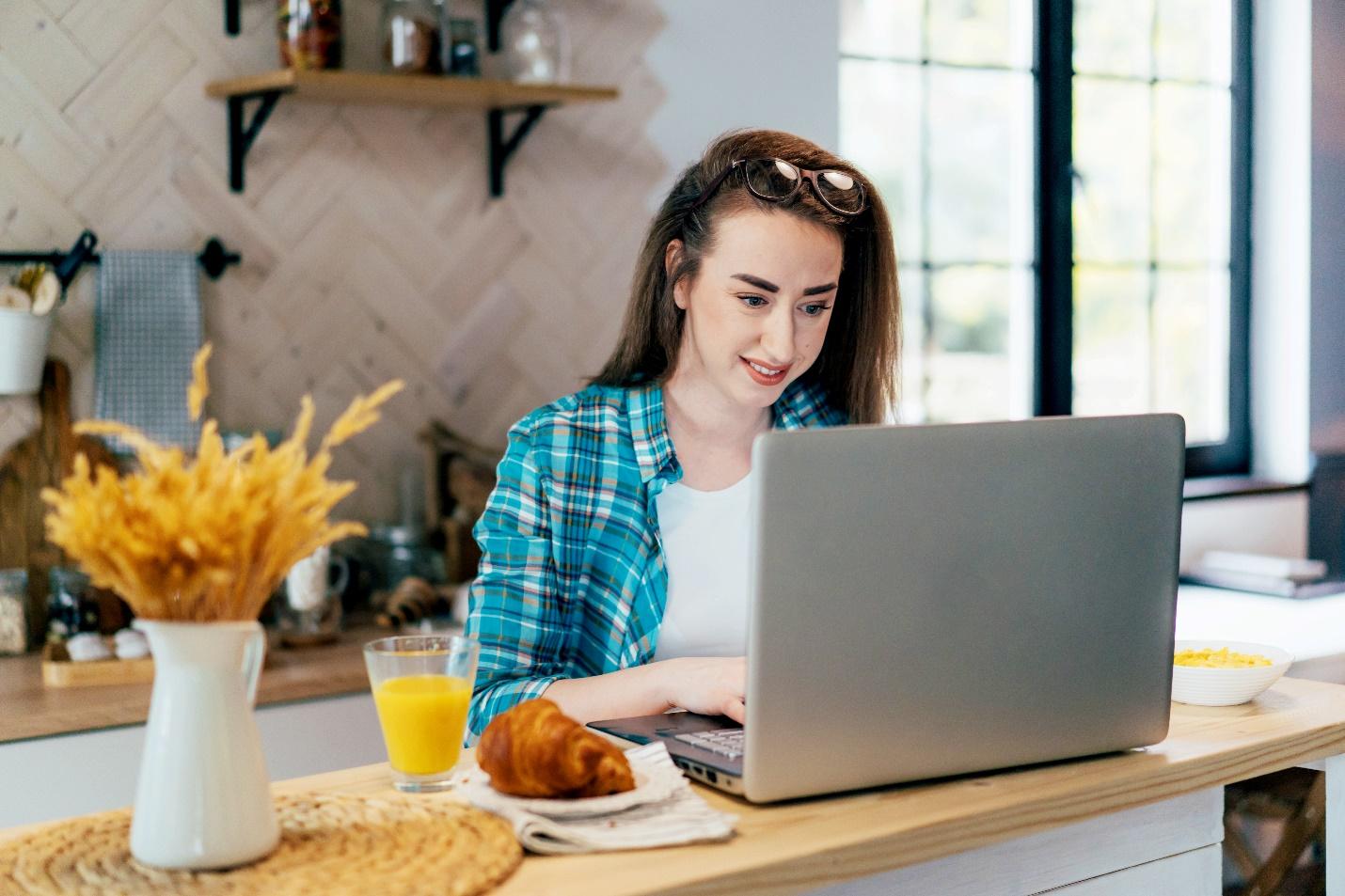 A casually dressed woman works at her laptop computer while eating breakfast at home.